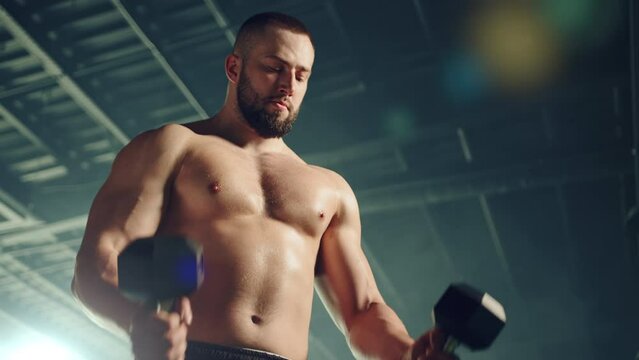 Medium close up of young athletic man with bare torso doing exercises with dumbbells. Handsome man is doing dumbbell biceps curl. Focus on arm workout. High quality 4k footage