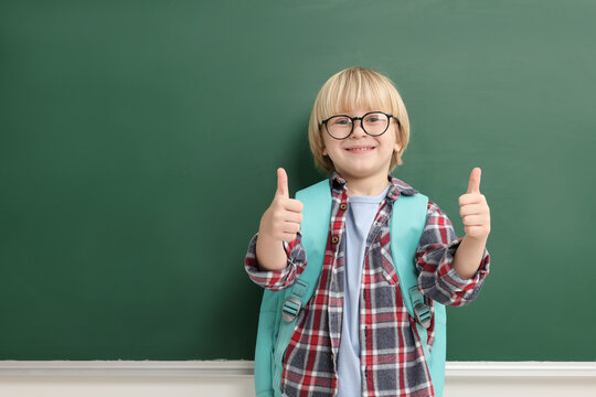 Happy little school child showing thumbs up near chalkboard. Space for text