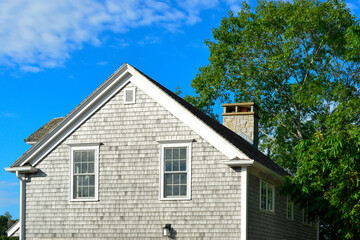 Two story grey colored vintage building with wood cedar shake siding, two double hung windows, and a chimney. There's a large lush green tree taller than the house. The trim on the building is white.