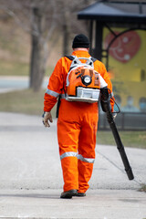 A man wearing a bright orange uniform suit with a mechanical battery operated leaf blower on his...