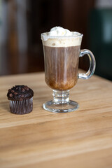 A clear glass mug of Irish coffee topped with whipped cream and a sugar rim. The tall glass is on a wooden bar at a pub. A chocolate muffin is on the wooden table. The cream is falling to the bottom.