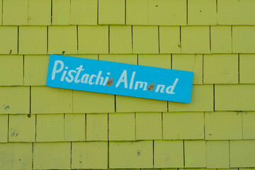 Obraz premium A vibrant lime green color external building wall made of cedar shakes. There's a colorful blue sign on a narrow board with white lettering spelling pistachio almond. The flavor of ice cream at a shop