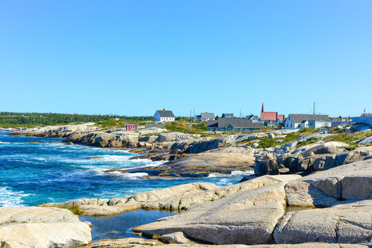 Scenic Peggy's Cove, Nova Scotia, is a quaint fishing village on the edge of a rocky coastline. The storage buildings are colored red, green, and blue color. The summer sky is blue with white clouds. 