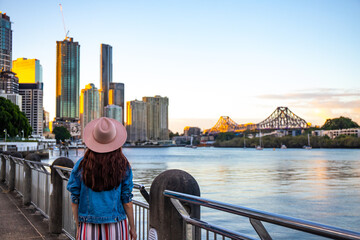 beautiful long hair woman in a hat watching the sunset over brisbane city; city reach boardwalk with amazing view of large skyscrapers by brisbane river, australia, holidays in brisbane - obrazy, fototapety, plakaty