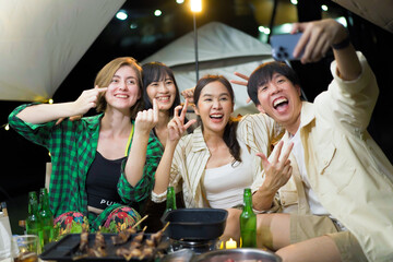 Group of diverse ethnicity young people enjoy eating and grilling bar-b-q together while camping in forest at night.