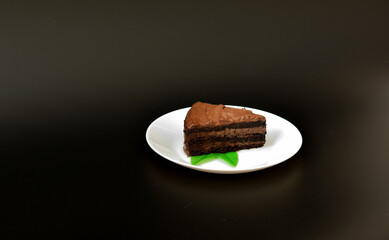A piece of biscuit chocolate cake decorated with mint leaves on a white ceramic plate on a black background.