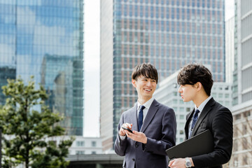 高層ビルに囲まれた都会の屋外で楽しそうに会話をする20代から30代のスーツのビジネスマンの男性2人