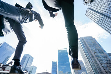 ビル群に囲まれたビジネス街でローアングルから撮影したジャンプとダッシュするスーツの2人の男性 - obrazy, fototapety, plakaty