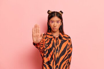 Young woman with funny hair style and stylish make-up stands on pink studio background, dressed in zebra design shirt, extends hand forward as stop sign, copy space, high quality photo