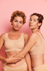 Two girlfriends posing inside the studio, curly one standing full face, her friend hugs her around the waist, both dressed in beige sport bras, friendship concept, copy space, high quality photo