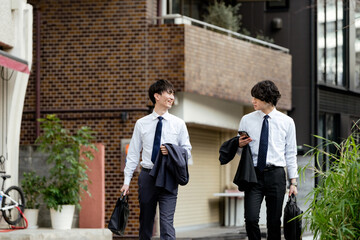 街をスーツのシャツ姿でスマートフォンを持ち会話しながら並んで歩く2人のビジネスマンの笑顔の男性
