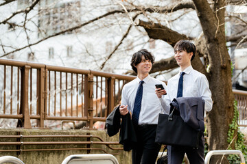 桜の緑道のある街並みをスマートフォンを持って会話しながら笑顔で並んで歩く2人の若いビジネスマンの男性 - obrazy, fototapety, plakaty