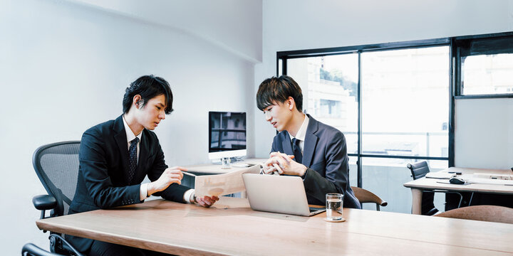 中小企業のオフィスの会議テーブルで打ち合わせをするビジネスマンの男性社員とインターンシップの男性