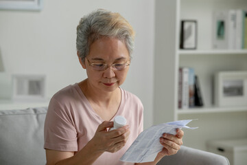 Asian senior woman sitting on sofa in her living room reading the information sheet of her prescribed medicine