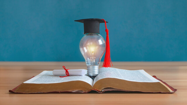 A Graduation cap and a graduation roll that tied with red ribbon rested on an open book on the table.Mortarboard with light bulb isolated on blue background.  It is the concept of education