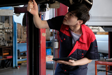 Professional young Asian male motor mechanic inspects undercarriage of electric car(EV) lifted by forklift jack for repair at garage, automotive maintenance service works industry occupation business.