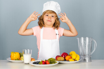 Fruits and vegetables for kids. Child chef cook prepares food on isolated grey studio background. Kids cooking. Teen boy with apron and chef hat preparing a healthy vegetables meal in the kitchen.