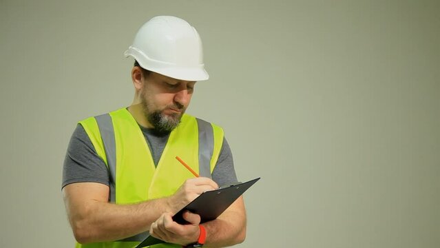 A male worker in a vest and a white construction helmet hat holds a folder with a clip for sheets of paper on a white background..
