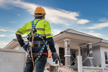 A construction worker stands holding an air gun or pneumatic nail gun and wearing a safety belt for...