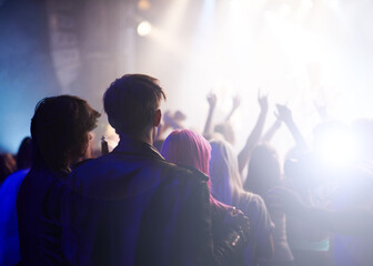 Fototapeta premium Dance, rock and friends in crowd at music festival from back, watching live band performance on stage. Audience in arena, lights and group of people at party, fans of musician at concert together.