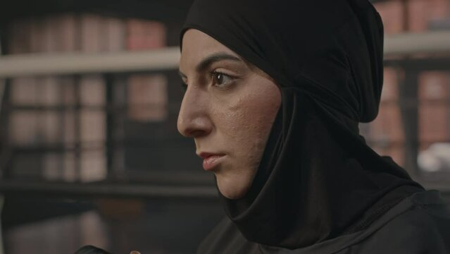Close up of confident young Muslim woman in hijab hitting punching bag with effort while having boxing training in dark club