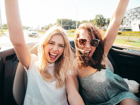 Portrait of two young beautiful and smiling hipster female in convertible car. Sexy carefree women driving. Positive models riding and having fun outdoors. Enjoying summer days. Cheerful and happy