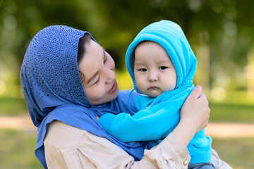 Cute young islamic kazakh woman hugging her little crying son, newborn boy. Yappy asian mother in hijab soothes, holding baby in hands outdoors. Family concept