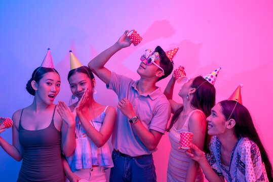 Five revelers holding cups and wearing party hats being silly and having a great time. People and plain background lit with pink and blue lights.