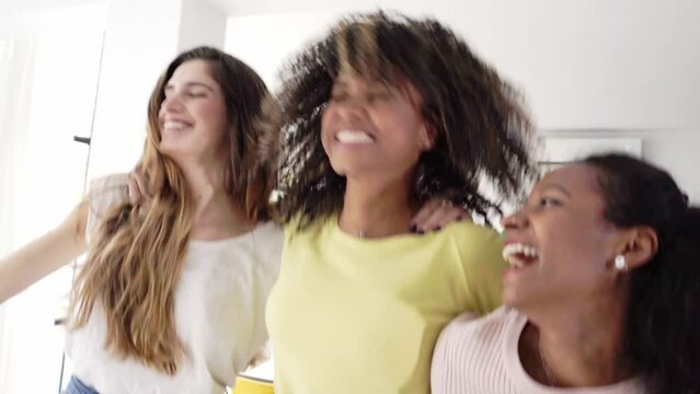 Three excited young friends dancing and singing at home. Cheerful multiracial girls enjoy together jumping hugging each other having fun. Happy leisurely people celebrating friendship. 