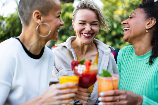 Three happy women friends having fun and cheering together with drinks hanging out in summer at restaurant