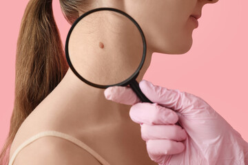 Dermatologist examining young woman's mole with magnifier on pink background, closeup