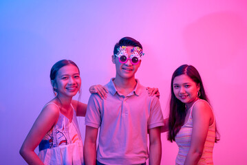 An grateful young man with two close female friends celebrating his birthday. Wearing funny novelty shades. Party scene lit with pink and blue neon colors.