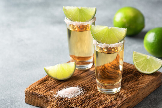 Mexican gold tequila in a shot glass with lime and salt on a wooden board, gray concrete background. Side view, selective focus.