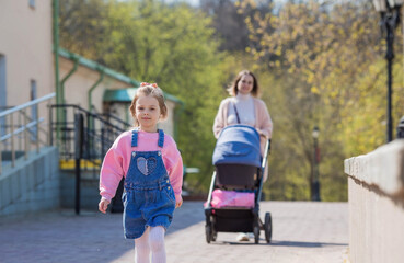 A beautiful little girl in pink clothes walks with her mother with her newborn brother in a stroller in the park in the fresh air. Family, children, motherhood.