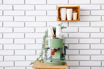 Modern coffee machine with paper cups and houseplant on table near white brick wall