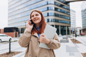 Young happy woman with laptop walking on the modern city street. Female worker using on mobile phone standing outdoors. Urban lifestyle concept