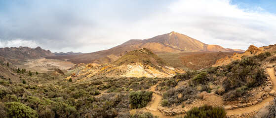Panoramic view of Teide National Park's rugged volcanic landscape, with iconic Pico del Teide and Pico Viejo peaks, surrounded by gravel fields and hiking trails with copy space and an overcast sky.