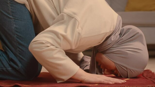 Side view stab shot oof young Muslim woman putting her head down on red carpet while praying at home