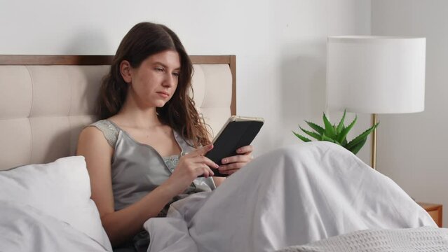 View of a young pleasant woman who lies in bed and looks at something on a digital tablet. A brunette woman woke up early in the morning and browses the social media feed on her tablet.