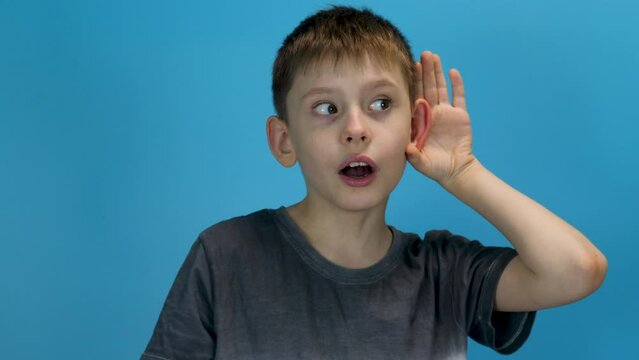 handsome curious caucasian boy 8 years old in casual clothes eavesdrops, studio shot blue background. concept of secrets, gossip, chatter. forbidden topics for children
