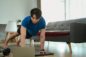  Motivated man in sportswear working out at home and watching fitness lessons online on laptop