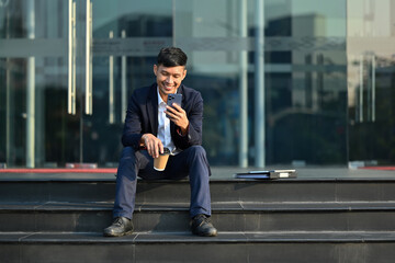 Attractive young businessman sitting on stairs and checking smartphone before going to the office