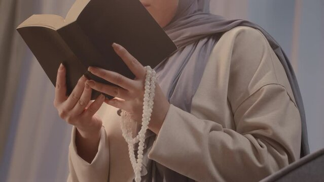 Low angle of young Muslim woman with rosary beads in hands reading holy book at home in evening