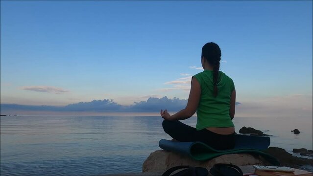 Woman in lotus position on a yoga mat meditating on a lakeshore at sunrise, seen from behind