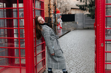 Fototapeta na wymiar A girl in a gray coat stands by a red telephone booth