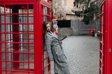 A girl in a gray coat stands by a red telephone booth