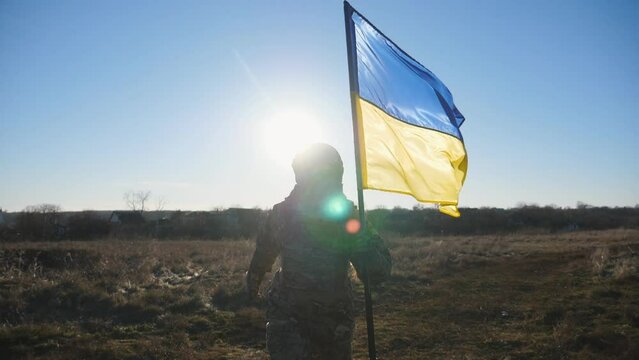 Female ukrainian army soldier running with lifted national banner on the field at sunny day. Woman in military uniform jogs with waving flag of Ukraine in honor of victory against russian aggression