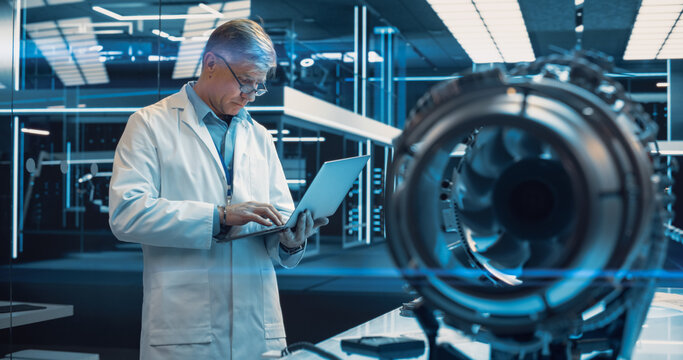 Adult Chief Engineer Using Laptop Computer, Analyzing and Researching How a Futuristic Turbofan Motor Works. Senior Manager Developing Innovative Technology in Industrial High Tech Facility