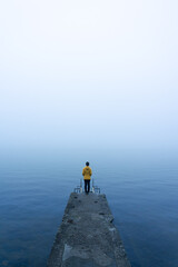 Fototapeta na wymiar Woman with a yellow jacket looking at the horizon on a pier on a very foggy day at Lake Sanabria. Zamora, Spain.