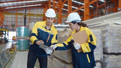 Two industrial worker man and woman in uniform using tablet checking quality and stock of metal sheet roof at manufacturing factory